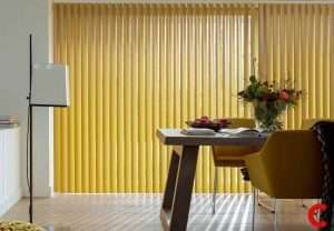 Mustard yellow vertical blinds behind a wooden dining table set with fruit and flowers