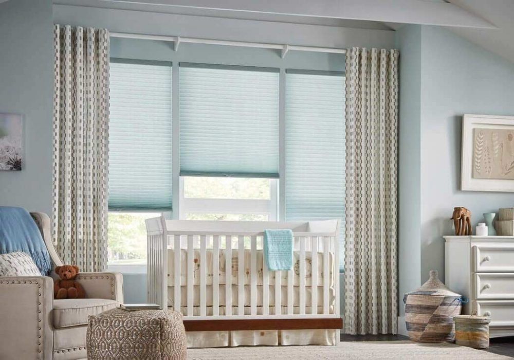 Patterned cream eyelet curtains beside pale blue cellular shades over a nursery window, white cot below