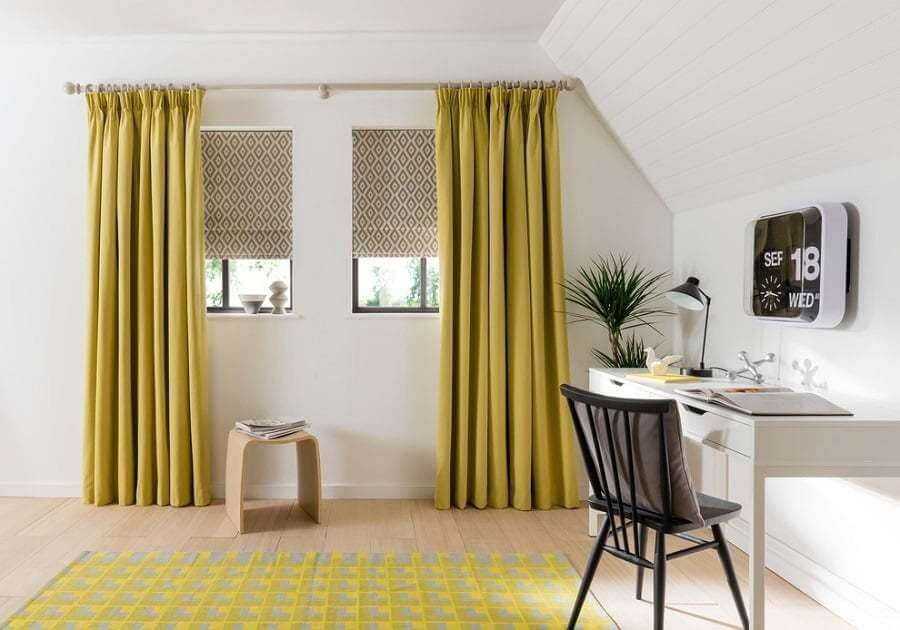 Mustard curtains on a wooden pole flanking two small windows fitted with patterned roman blinds in an attic study