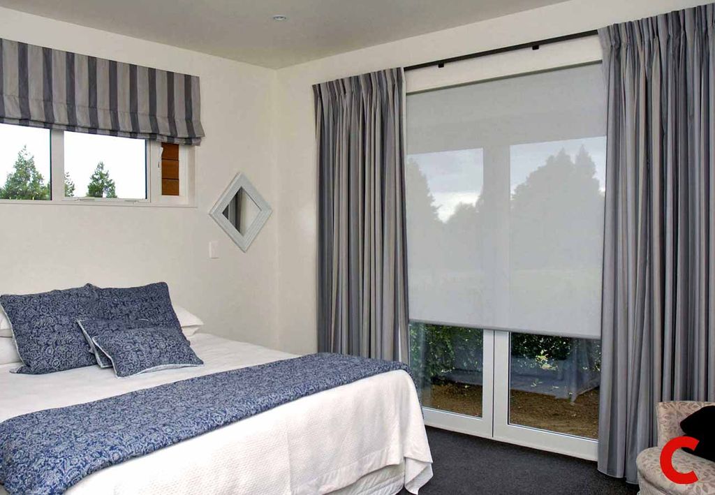 Grey curtains framing a patio door with a white roller blind half down, and a striped roman blind above the bed