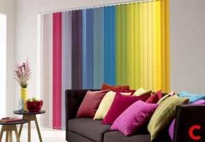 Vertical blinds in rainbow-coloured bands across a window above a brown sofa with matching bright cushions