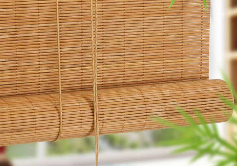 Close crop of a honey bamboo roll-up blind with its lift cords and rolled bundle, foliage at the edge