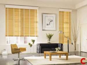 Honey-toned wooden venetian blinds, slats tilted open, on two windows behind a black sofa