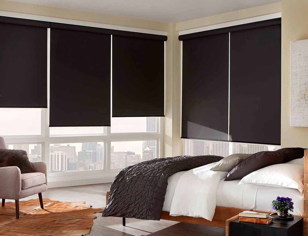 Black blackout roller blinds lowered over bedroom corner windows, city towers still bright below the hem