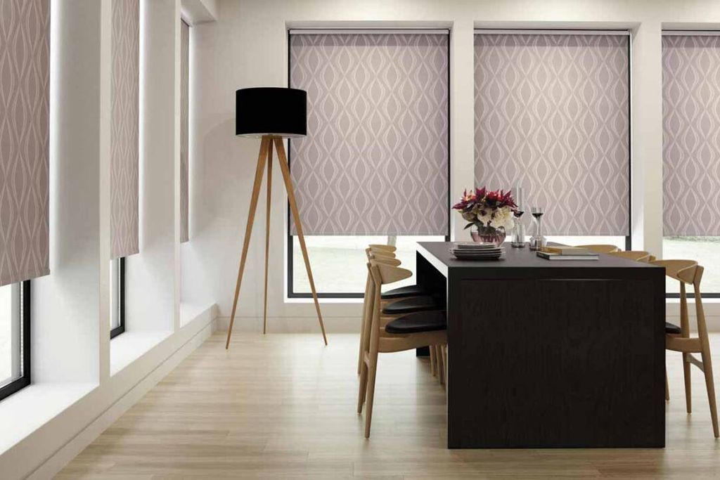 Dusky pink patterned roller blinds fully lowered across a dining room's tall windows beside a dark wood table