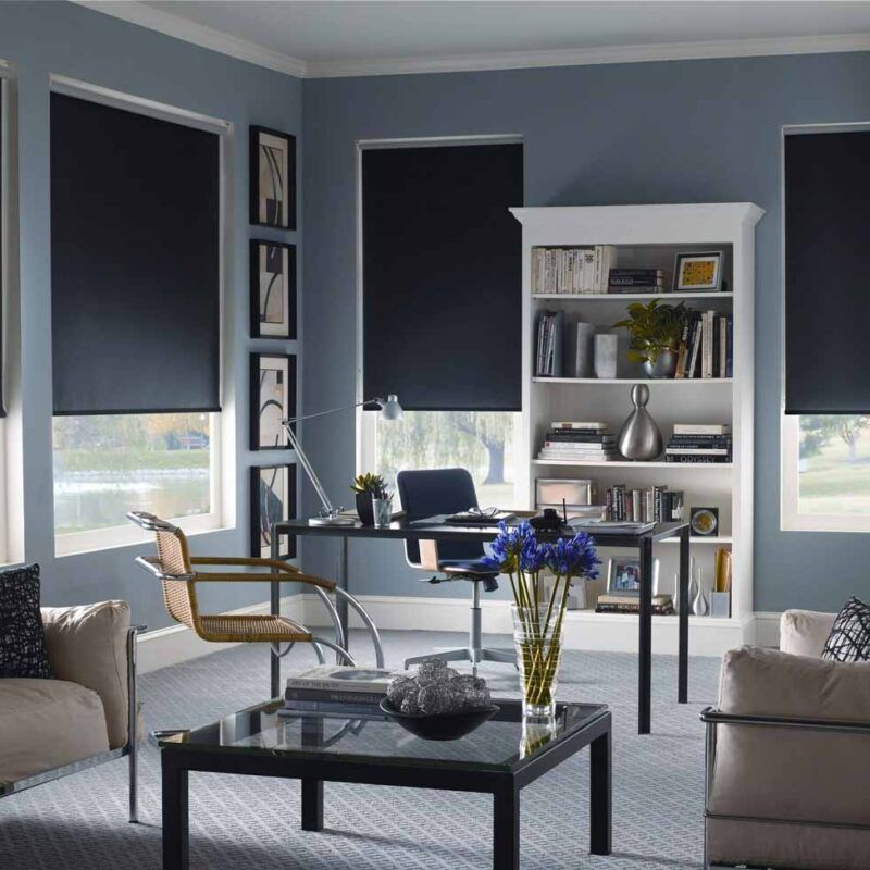 Navy roller blinds three-quarters lowered at three windows of a blue-grey home office with a white bookcase