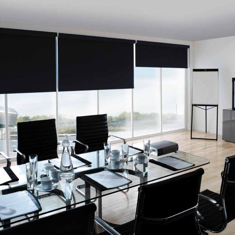 Black roller blinds half lowered along a boardroom's glazed wall, above a glass table and black leather chairs