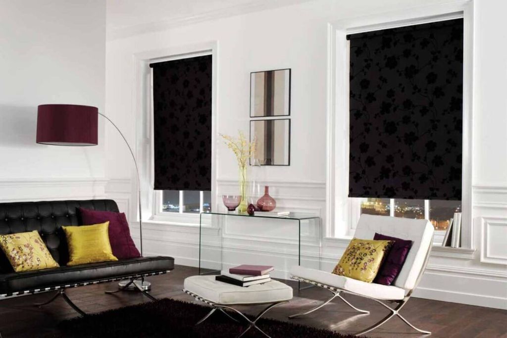 Black floral-print roller blinds lowered over two tall sash windows in a white panelled living room at night