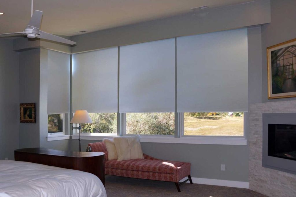 Pale grey roller blinds lowered almost fully across a wide bank of bedroom windows beside a stone fireplace
