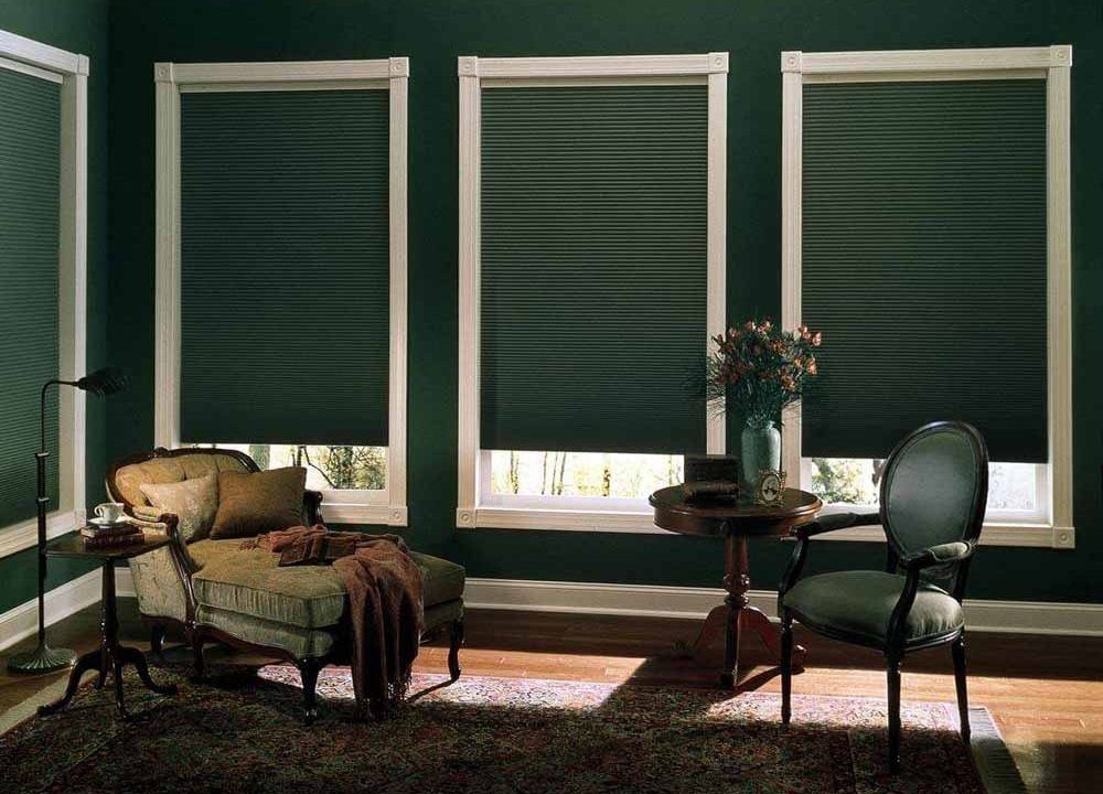 Dark green pleated blinds lowered near the sill on tall windows in a green sitting room with an antique chaise