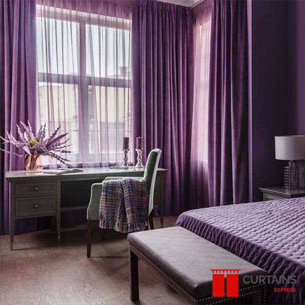 Purple floor-length curtains framing a corner bedroom window, with a matching sheer drawn across the glass