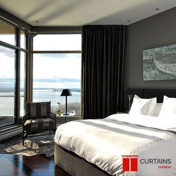A black floor-to-ceiling curtain on a ceiling track beside a corner window wall in a grey bedroom with a lake view