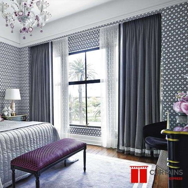 Grey pleated curtains layered over white sheers at a tall bedroom window, against navy geometric wallpaper
