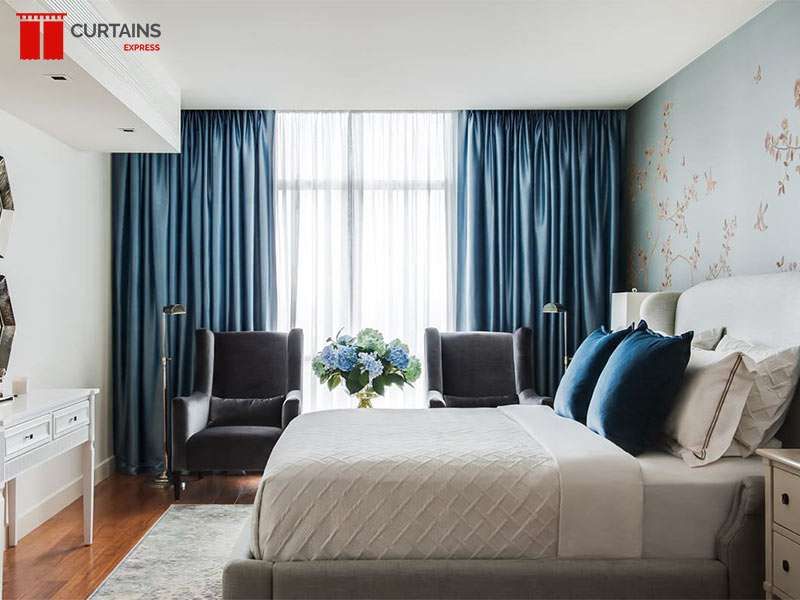 Teal satin pleated curtains either side of a tall bedroom window with white sheers drawn across the glass