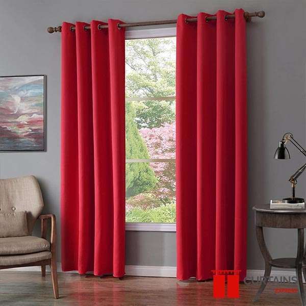 Red eyelet curtains on a wooden pole framing a window that looks onto a green garden, in a grey-walled room