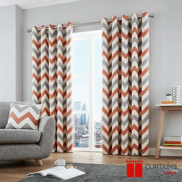 Orange and grey chevron eyelet curtains on a chrome pole at a full-height city window, beside a grey sofa