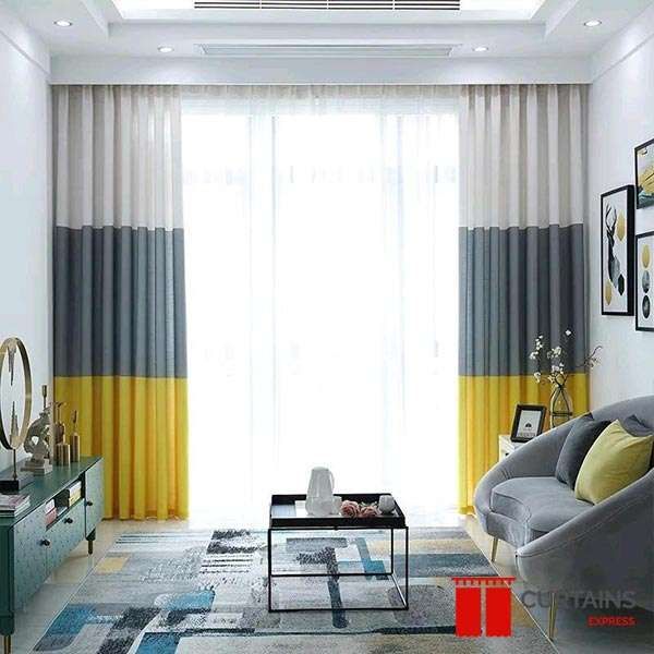 Cream, grey and yellow colour-blocked curtains on a ceiling track across a living room window, sheers behind