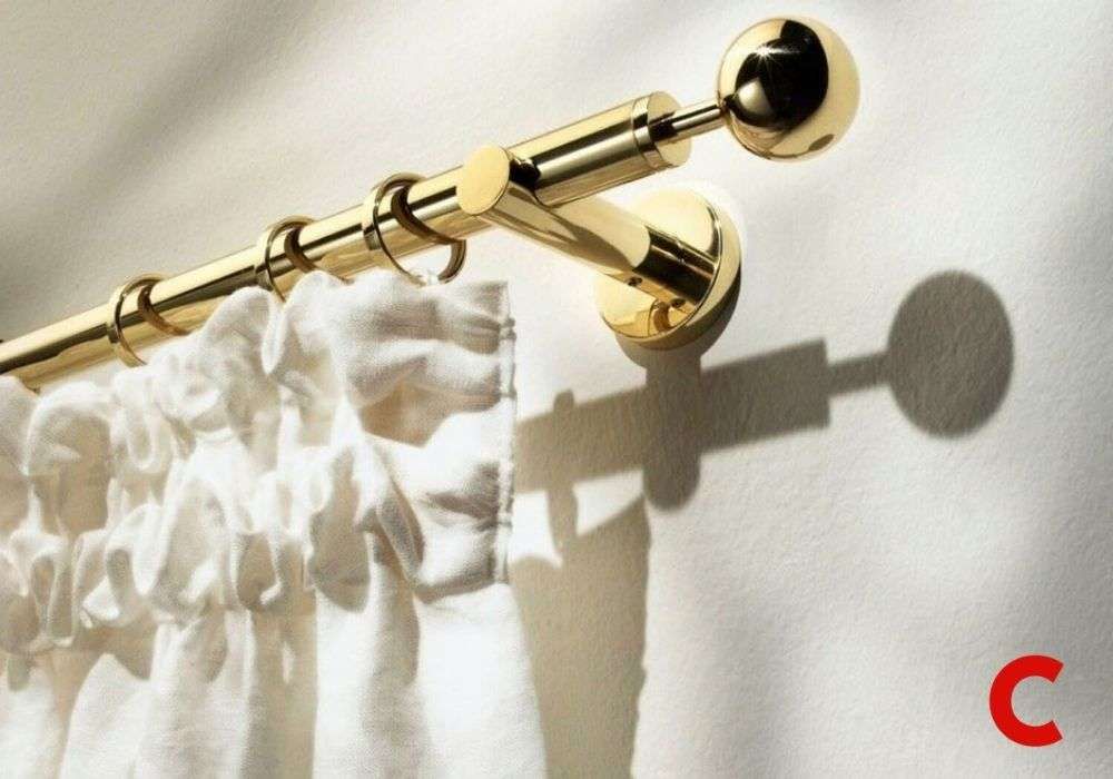 Polished brass curtain pole with a ball finial on a wall bracket, holding a gathered white curtain in bright sun