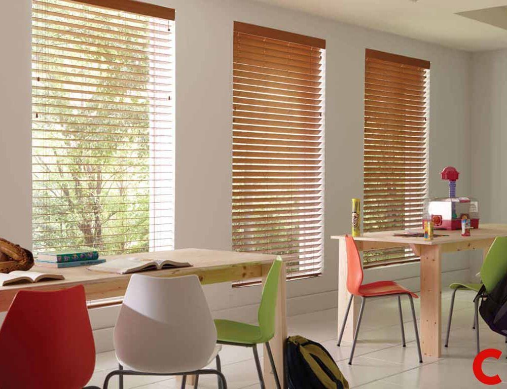 Mid-brown wooden venetian blinds on three tall windows of a bright classroom with pine tables and coloured chairs