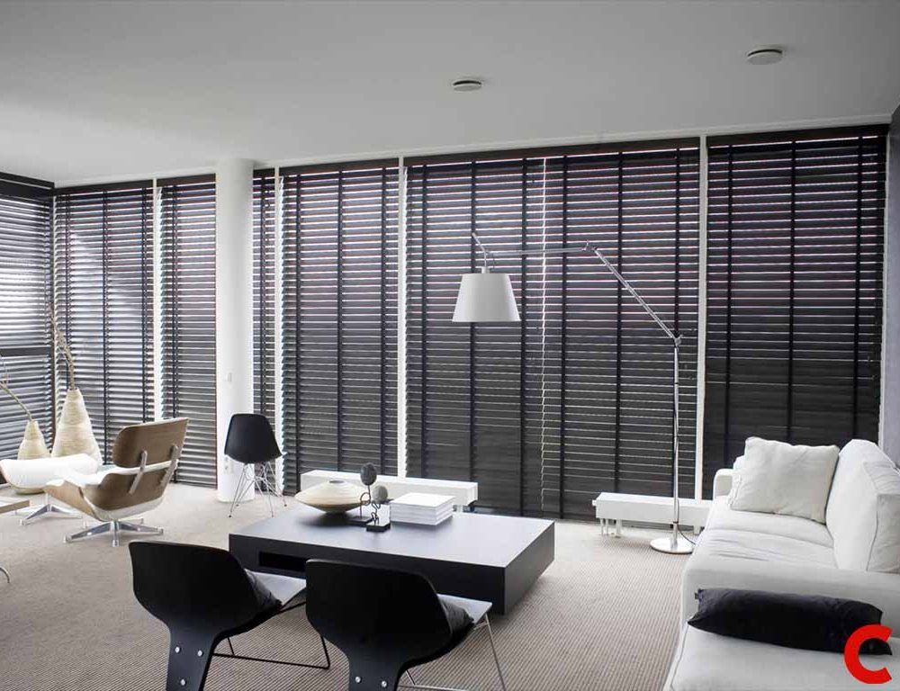 Wide-slat dark wooden venetian blinds covering floor-to-ceiling windows around a white minimalist lounge