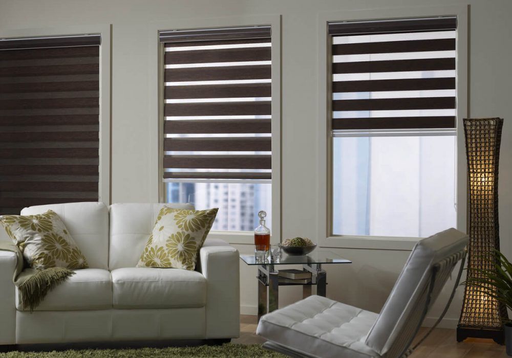 Dark brown zebra blinds set at three different heights on tall windows above a city view, white leather sofa