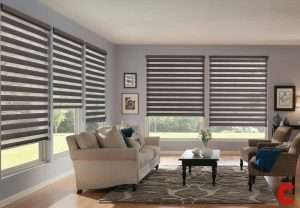 Grey-brown zebra blinds across three windows of a lilac-grey living room with a cream sofa and armchair