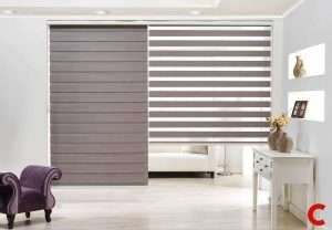 Two grey zebra blinds side by side, the left closed and the right open, beside a purple velvet armchair