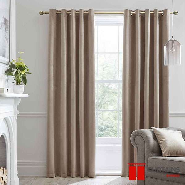 Taupe eyelet curtains on a brass pole framing a sash window, beside a white fireplace and grey buttoned sofa