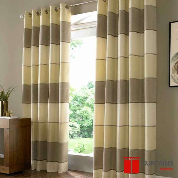 Cream, taupe and grey horizontally striped eyelet curtains drawn back from a window above a wooden sideboard