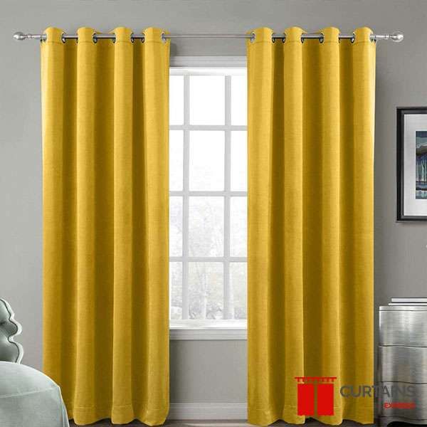 Mustard yellow eyelet curtains on a chrome pole either side of a small bedroom window in a grey room