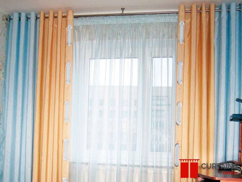 Pale blue and peach eyelet curtains layered over a white sheer at an apartment window facing another block