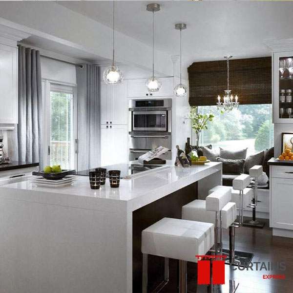 Grey full-length curtains at a garden door and a dark woven roman shade over the window seat in a white kitchen