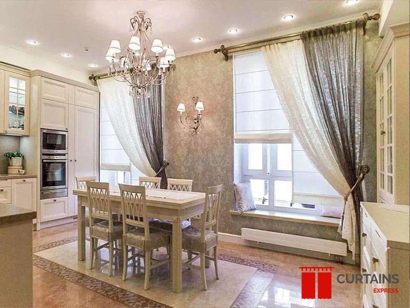 Taupe curtains and white lace sheers hooked back over a white roman blind at a kitchen-diner window seat