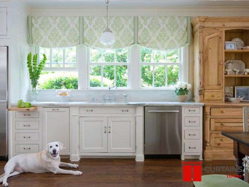 Three green damask roman shades raised above the windows over a white kitchen sink, with a pine dresser alongside