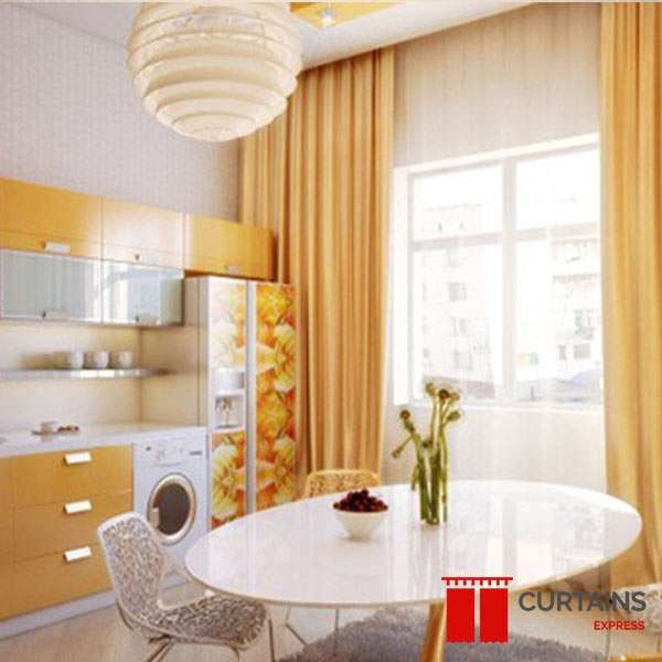 Golden yellow floor-length curtains framing a kitchen window beside an oval white dining table and yellow gloss units