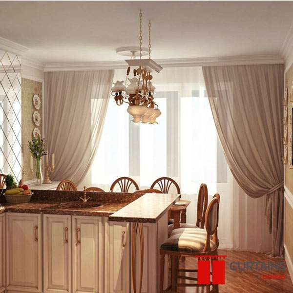 Mocha curtain panels tied back over white sheers at a kitchen-diner window, framing a cream island and chandelier