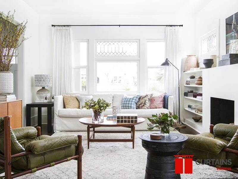 White pleated curtains stacked either side of a black rod spanning three windows above a white sofa
