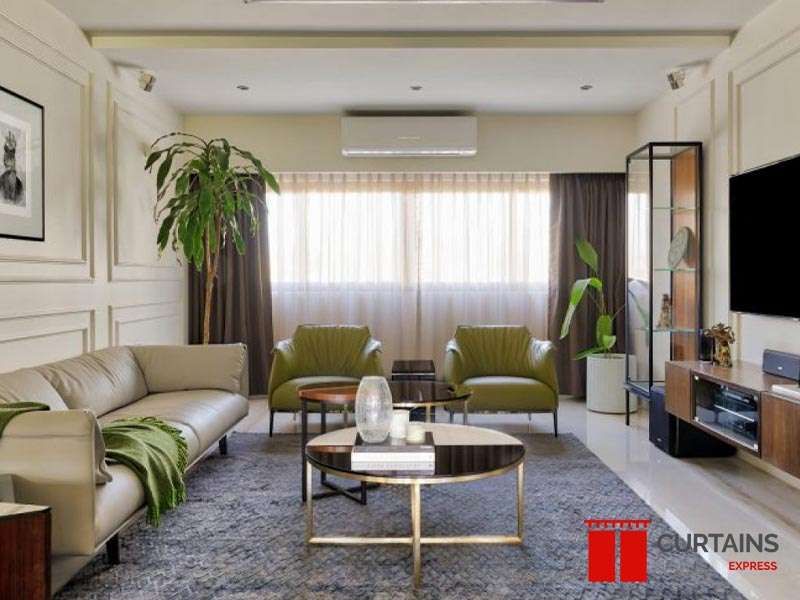 Dark brown curtains drawn back over a full-width white sheer in a living room with olive armchairs and a wall TV