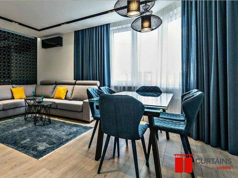 Teal floor-to-ceiling curtains framing a corner window with a white sheer drawn across, in a grey open-plan room