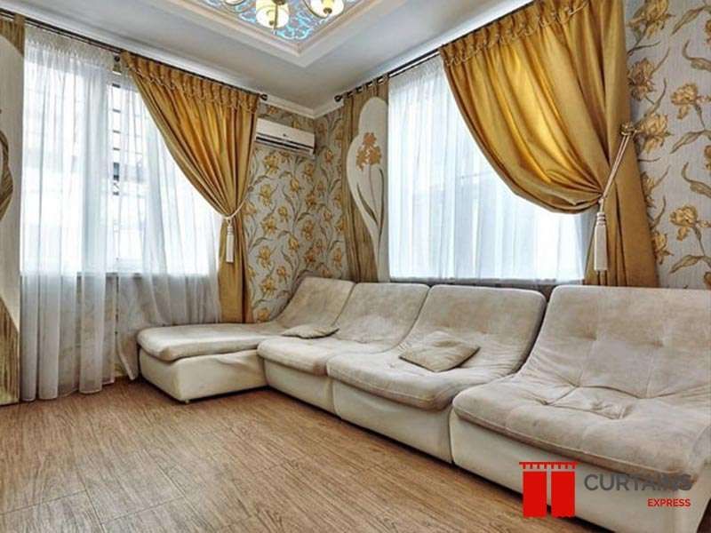 Gold satin curtains held by tassel tiebacks over white sheers at two windows in a floral-wallpapered lounge