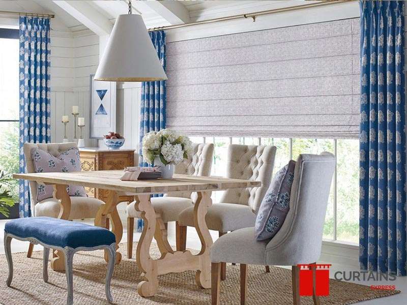 Blue block-print curtains at the windows and a grey roman blind lowered over the centre pane above a rustic table