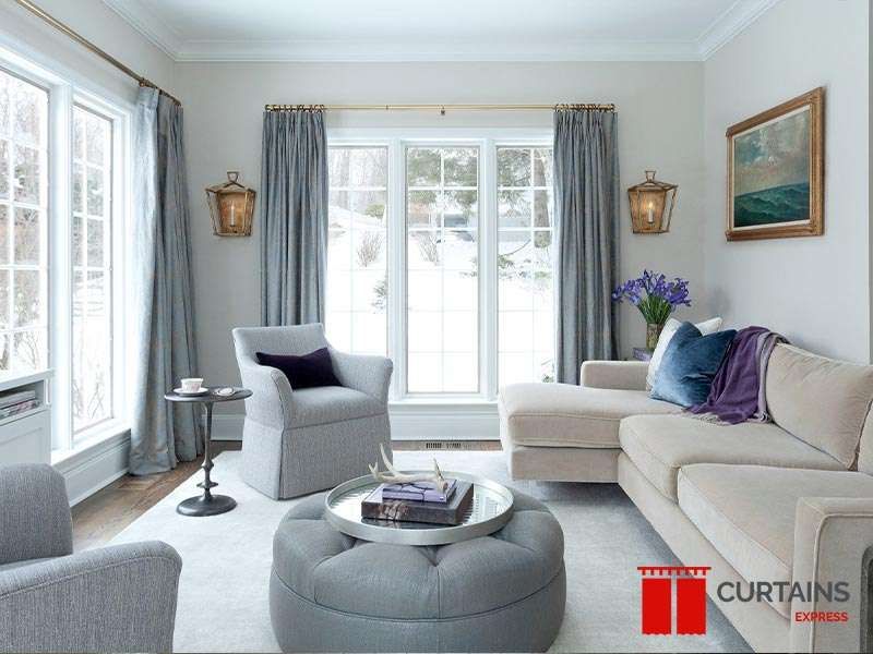 Pale blue-grey pleated curtains on brass rods at two windows, snow outside, above a cream sectional and ottoman