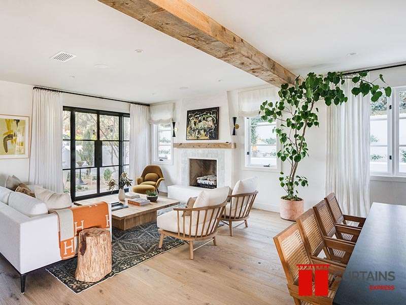 White floor-length curtains at steel-framed windows and cream roman shades flanking a fireplace in a beamed room