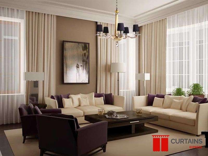 Beige floor-to-ceiling curtains layered over white sheers on two walls of a lounge with aubergine armchairs