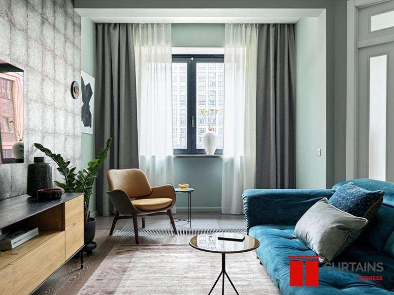 Grey curtains and white sheers framing a window recess with a city view, beside a teal velvet sofa