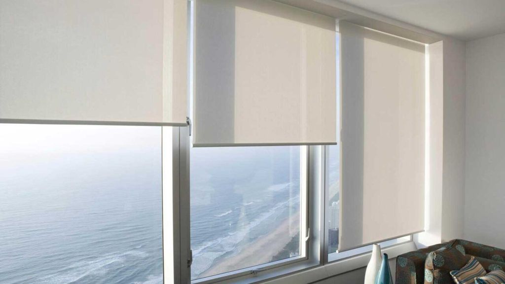 Cream roller blinds lowered to different heights across high-rise windows overlooking a city