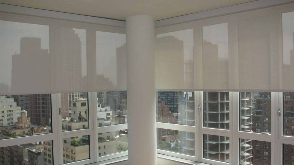 Translucent white roller blinds half lowered across corner windows, city towers showing through the screen fabric
