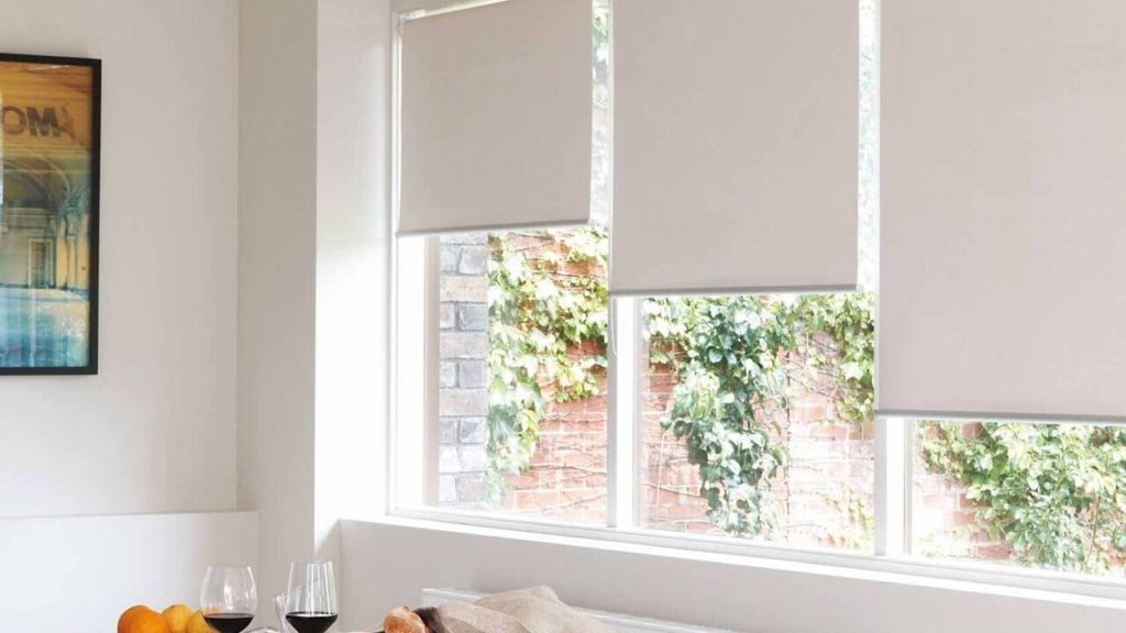 Three greige roller blinds part-lowered over a run of windows looking onto an ivy-covered brick wall