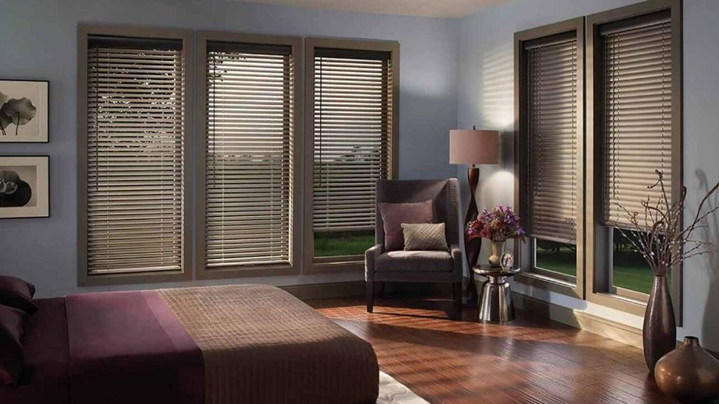 Bronze wide-slat wooden venetian blinds across five bedroom windows, with an armchair and lamp in the corner