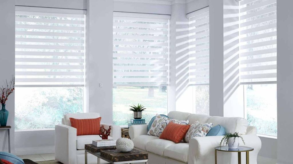 Sheer white shades with horizontal fabric vanes across corner windows in a bright living room with coral cushions